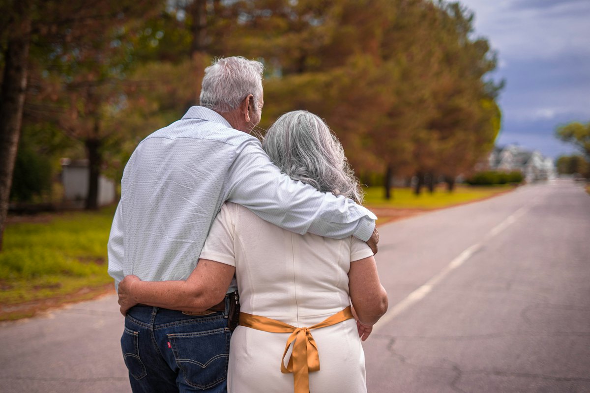 Casal de cabelos grisalhos caminhando abraçado por uma estrada arborizada